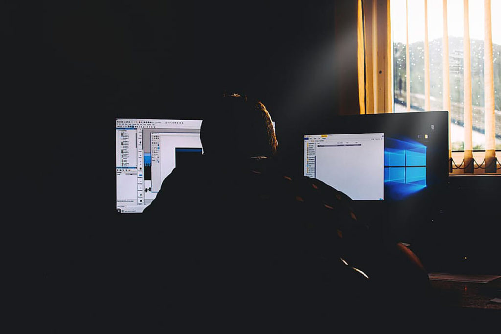 Человек сидит перед компьютерными мониторами у окна в темной комнате